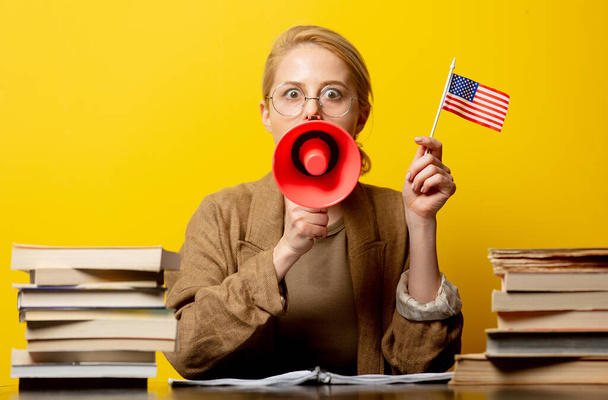 Stijl blonde vrouw in zitten aan tafel met USA vlag en megafoon met boeken rond op gele achtergrond - Foto, afbeelding