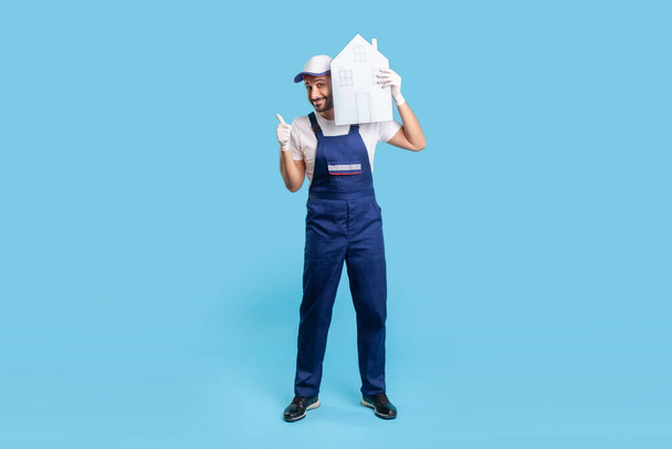 Full length happy labourer in uniform holding paper house on shoulder and showing thumbs up. Mover guy recommending help in relocation, loader services. indoor studio shot isolated on blue background - Foto, immagini