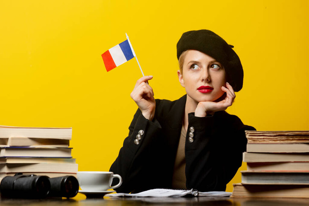 Bere takmış zarif sarışın kadın masada Fransız bayrağı ve sarı arka planda kitapları ile oturuyor. - Fotoğraf, Görsel