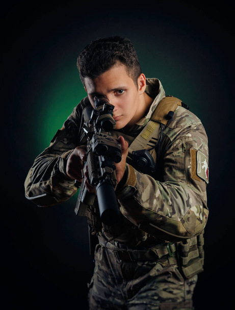 ein brutaler Kerl in militärischen Airsoft-Overalls posiert mit einer Waffe in den Händen vor dunklem Hintergrund - Foto, Bild
