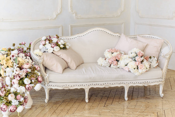 Canapé beige clair de couleur délicate à côté d'un grand nombre de fleurs, roses et pivoines - Photo, image