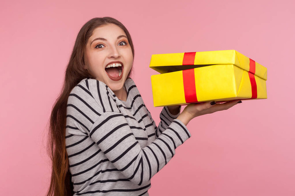 Портрет счастливой взволнованной женщины в полосатой толстовке, с удивлением выглядящей и радостно кричащей, открывающей подарочную коробку, распаковывающей лучший подарок, праздничный сюрприз. крытая студия съемки изолированы на розовом фоне
 - Фото, изображение
