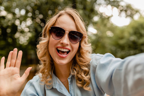 attractive blond smiling woman walking in park in summer outfit blue shirt taking selfie photo on phone, wearing elegant sunglasses, street fashion style - Fotografie, Obrázek