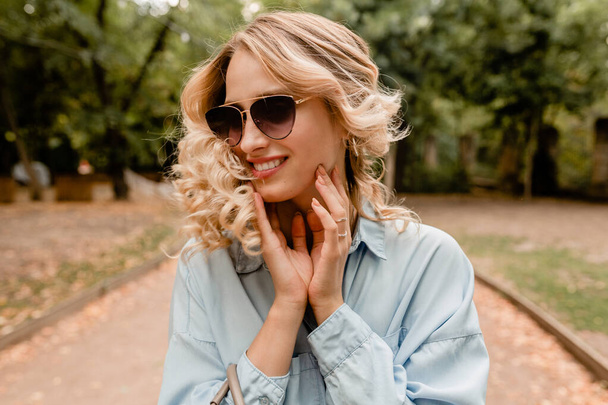 attractive blond smiling candid woman walking in park in summer outfit blue shirt, wearing elegant sunglasses, street fashion style, happy mood, waving curly hair - Photo, Image