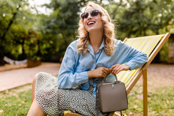 魅力的なブロンドの笑顔の女性は、エレガントなサングラスを身に着けて、夏の衣装ブルーシャツでデッキチェアに座って、財布、ストリートファッションスタイルのアクセサリー - 写真・画像