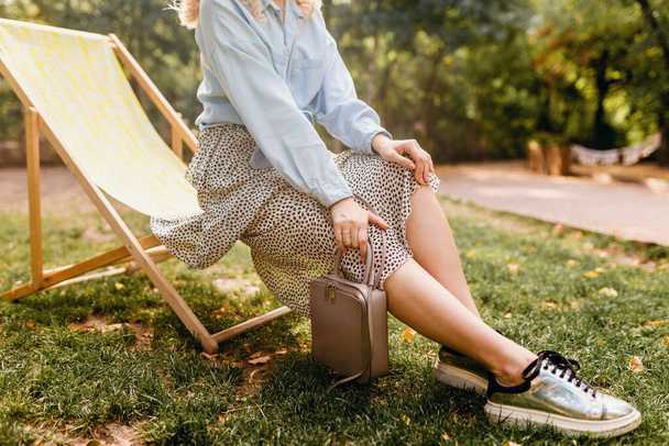 close-up legs in silver sneakers shoes, fashion autumn trend footwear, attractive woman sitting in deck chair in stylish outfit blue shirt and skirt, holding purse, street style accessories - Foto, Imagem