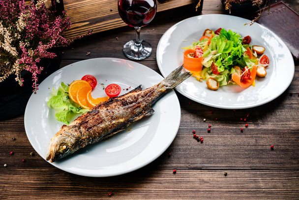 Grilled sea bass on the wooden board, seabass with salad, two dishes on the table - Foto, Bild