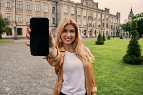 Excited young woman in casual clothes showing display of modern smartphone enjoying walk in street of city looking at camera - Фото, изображение
