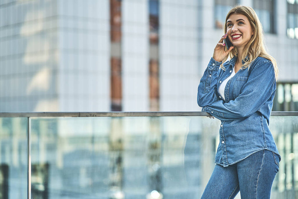 Young stylish girl wearing on denim clothing spending free time outdoors standing talking on smartphone with friend looking aside laughing cheerful close-up - Photo, image