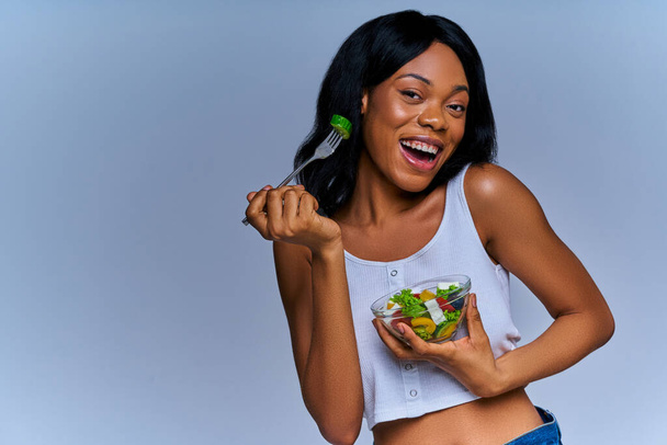 Pleased girl in white top enjoys mixed salad from glass bowl - Fotografie, Obrázek