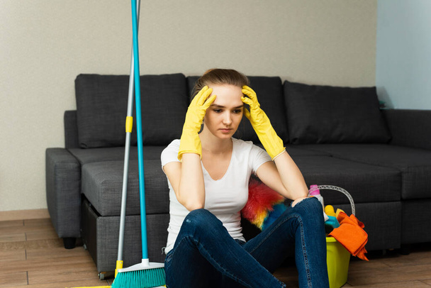 Uma mulher bonita em luvas de borracha ao lado de um casaco e produtos de limpeza fica com raiva de um monte de tarefas domésticas. Problemas de repartição das tarefas domésticas. - Foto, Imagem