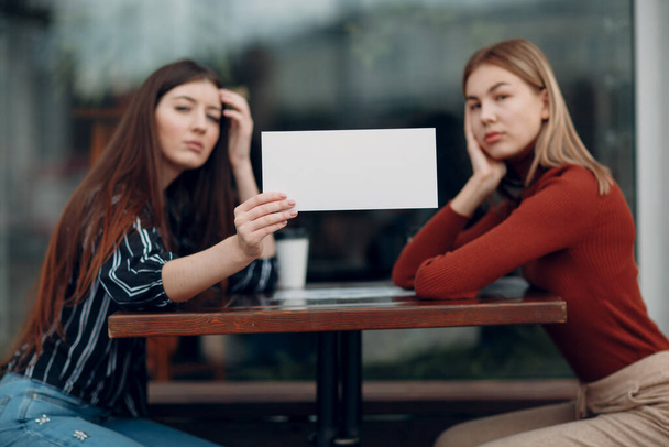 Woman holding white paper in hand. Two women talking at table in street cafe. White blank template sheet with empty space. - Foto, imagen