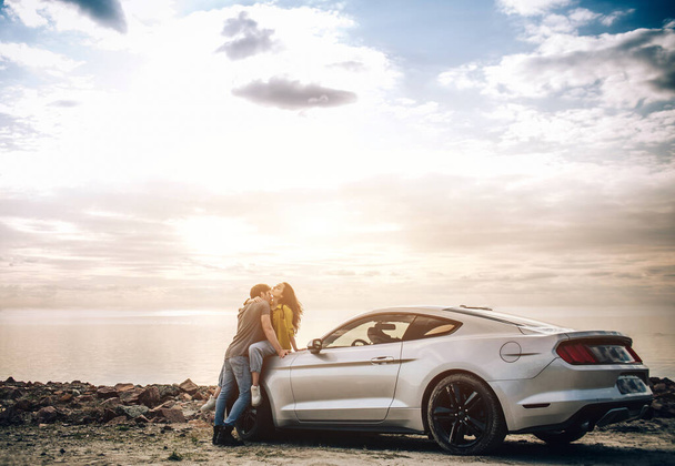 Romantic young couple sharing a special moment while outdoors. Young couple in love on a road trip. Couple embracing each other while sitting on hood of their car in nature. - 写真・画像