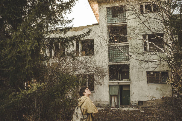 A boy inspects an unfamiliar place. A man in a protective jacket stands in front of an abandoned building. Large overgrown airport building. wanderer in the post-apocalyptic world - Photo, Image