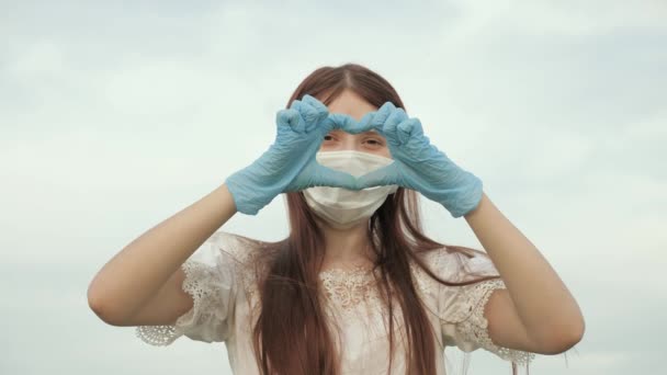mujer joven ama a los médicos. pandemia, covid 19. chica en una máscara protectora y guantes médicos muestra símbolo del corazón con sus manos. doctora mostrando los dedos del corazón y el amor por los pacientes
. - Imágenes, Vídeo