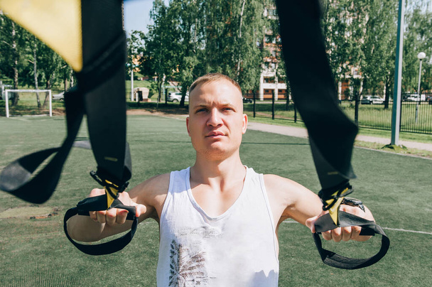 Athlete guy does pectoral muscle wiring, trains in summer city, holds loops his hands, sports straps for push-ups pull-ups, fitness, workout. Sportswear T-shirt. Background trees, sand sports ground - Foto, Imagen