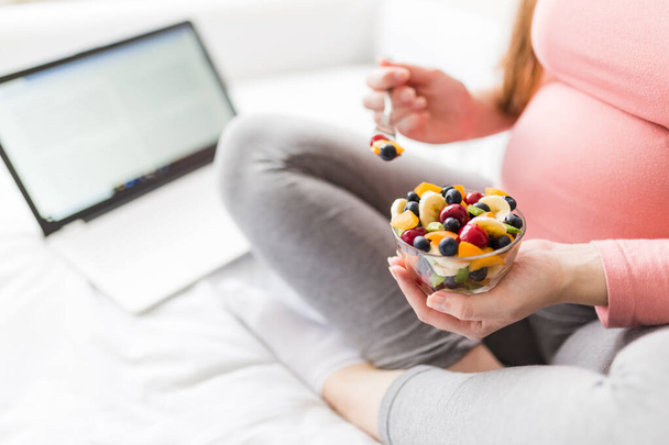 Young female eating fresh fruit salad and using laptop while sitting on bed, healthy breakfast concept - Valokuva, kuva