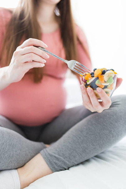 Close up of pregnant woman sitting on bed and eating fruit salad. Healthy meal concept. - Фото, изображение