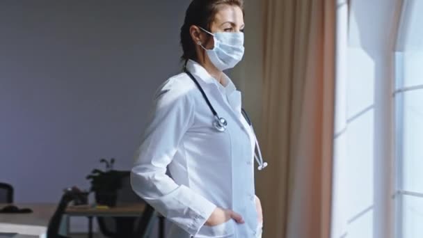 In the hospital mature doctor woman with a protective mask have a break time she came to the panoramic window and thinking - Materiał filmowy, wideo