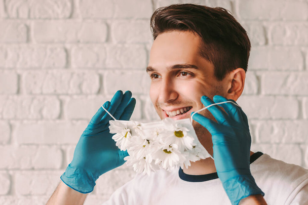 Portrait of young male doctor in protective gloves wearing or take off medical face mask with daisy flowers. Happy man put on summer protective face mask with flowers and smiling. COVID-19 quarantine - Foto, immagini