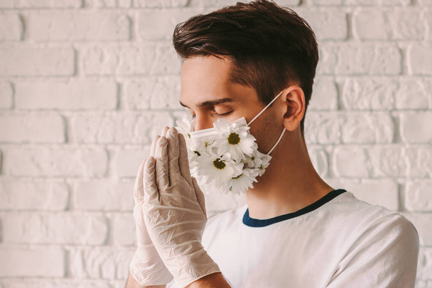 Portrait young faithful male doctor in medical face mask with daisy flowers praying to God with hope of coronavirus COVID-19 help. Religious man christian wearing protective mask with praying hands - Fotografie, Obrázek