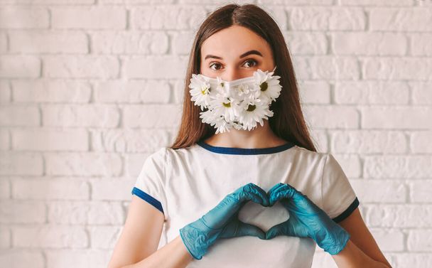 Portrait happy beautiful girl wear medical face mask with flowers, show love symbol with hands in protective gloves. Young woman doctor in summer protective mask gesturing heart shape sign by fingers - Fotoğraf, Görsel