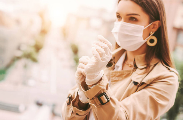 Beautiful young stylish woman in protective face mask put on medical latex gloves outdoors. Trendy elegant hipster girl in medical face mask wear protective gloves on city street. COVID-19 protection - Foto, afbeelding