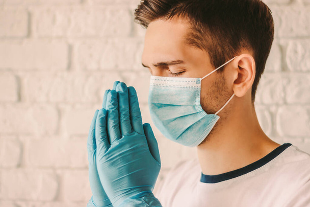 Young man patient in medical face mask and protective gloves on hands praying at home. Hope for help against coronavirus COVID-19. Surgeon doctor prayer in protective mask on face and medical gloves - Foto, Imagem