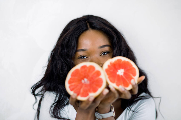 African American woman with long dark hair, smiling for camera and hiding half of her face with juicy grapefruit slices, standing against white background. Beauty portrait, copy space - Fotó, kép