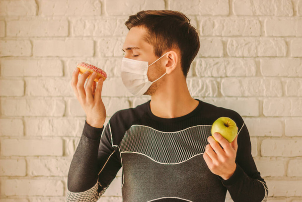 Man fitness trainer in medical face mask holds donut and apple in hands. Sports man in protective face mask choosing between healthy food or sugar snack. Dieting, home quarantine, coronavirus COVID-19 - Valokuva, kuva
