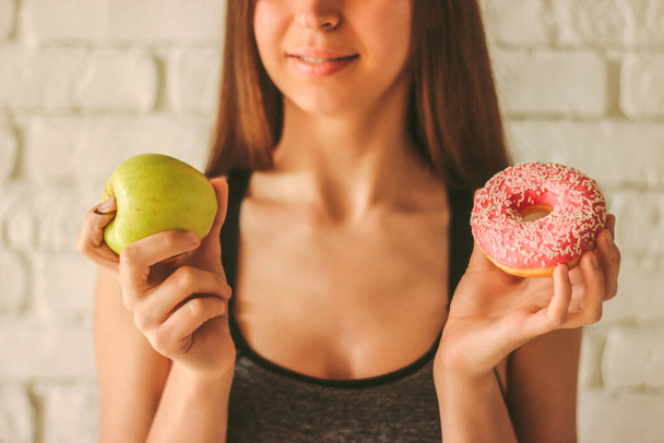 Closeup young sexy fitness girl holding apple and donut in hands. Beautiful slim sports woman choosing between healthy fruit or sweet donut temptation. Healthy lifestyle, fitness dieting, weightloss - Foto, immagini