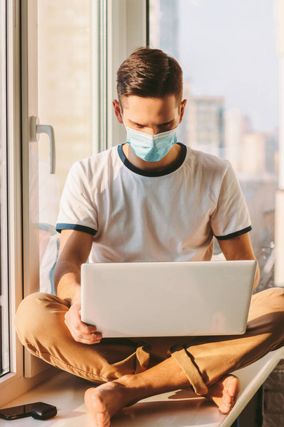 Young man in medical protective mask on face sitting on windowsill with laptop on laps. Student freelancer working at home. Distance job with computer. Business from home. Modern internet blogger - Foto, immagini