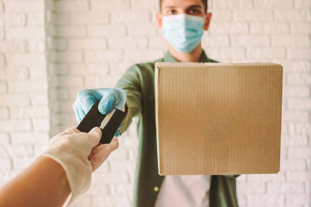 Happy hipster delivery man in medical face mask hold cardboard box in hand, checkout customer. POV customer in rubber protective gloves receive shipment box from courier. COVID-19 quarantine delivery - Фото, изображение