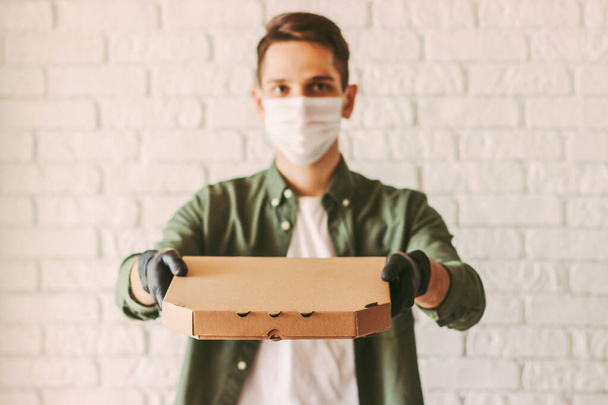 Hombre de entrega feliz en máscara facial médica y guantes sostienen caja de cartón de sabrosa comida rápida italiana en las manos. Hipster chico mensajero en máscara protectora llevar caja de pizza. Servicio de entrega de alimentos en cuarentena
 - Foto, imagen