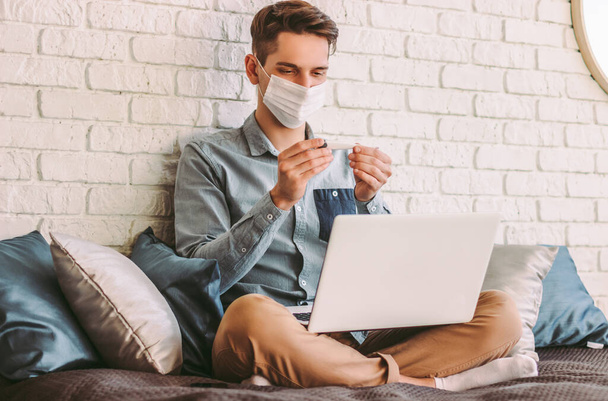 Sick hipster man student in medical face mask look at digital thermometer in hands, work at home. Young businessman freelancer in protective mask use electronic thermometer. Measure body temperature - Foto, Imagen
