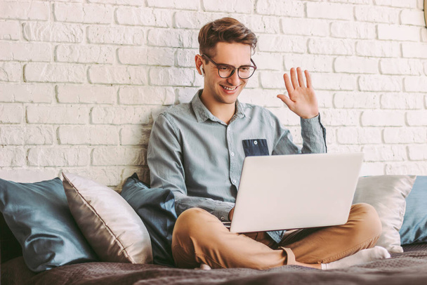 Stylish young hipster man student in glasses waving hand to laptop while talking online video call and sitting on couch. Happy businessman freelancer having web meeting, conference. Work at home - Foto, Bild