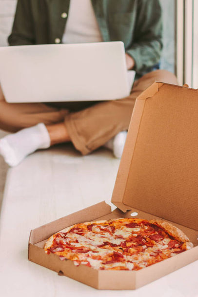 Closeup of cardboard box with fresh tasty cheese pizza on wooden windowsill. Hipster man freelancer working with laptop on background. Delicious italian fast food lunch for businessman work from home - Foto, imagen