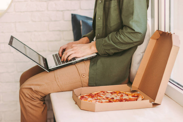 Closeup stylish hipster man typing laptop keyboard, eat delicious pizza, sit on windowsill. Busy businessman freelancer work remotely from home office and eat takeaway italian fast food. Lunch break - Фото, зображення