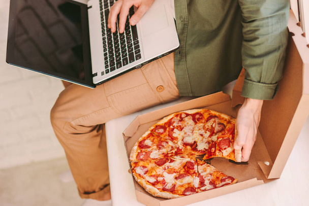 Top view nälkäinen liikemies syö maukasta italialaista pizzaa, käyttäen kannettavaa tietokonetta etätyön päässä kotitoimistosta. Tyylikäs hipster mies kirjoittamalla tietokoneen näppäimistö, syödä takeaway pikaruokaa kodikas koti työpaikalla - Valokuva, kuva