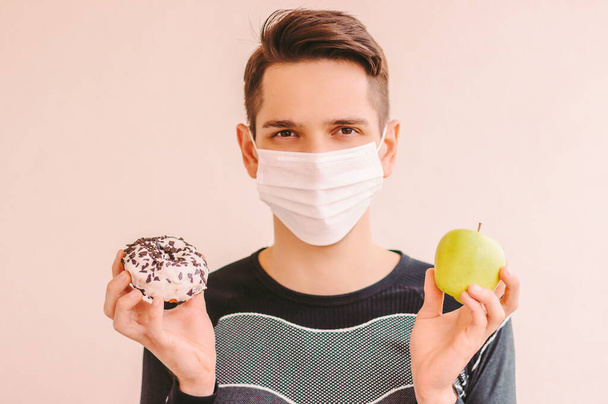 Happy sports man in medical face mask holding sweet donut and fresh apple in hands. Young fitness man in protective face mask struggling between healthy and unhealthy food. COVID-19 quarantine diet - Foto, Imagem