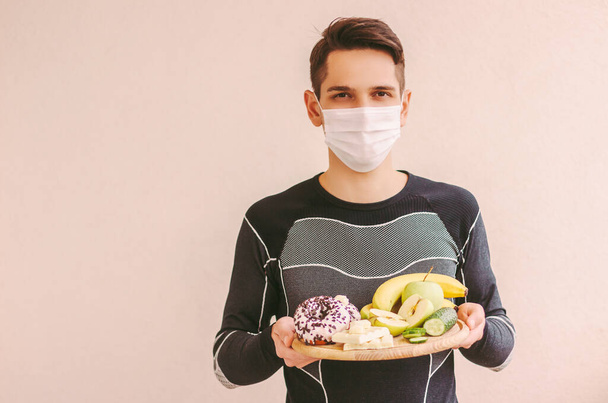 Young fitness man in protective mask with wooden board in hands full of fruits, vegetable, sweet donut and chocolate. Happy sports man in medical face mask choose food for diet. Coronavirus quarantine - Valokuva, kuva