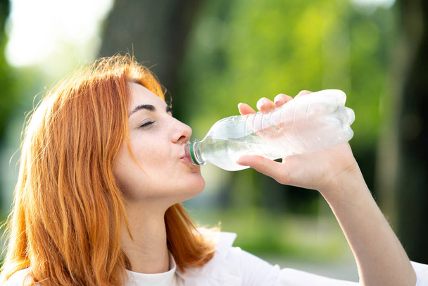 Junge durstige rothaarige Frau trinkt Wasser aus einer Flasche im Sommerpark. - Foto, Bild