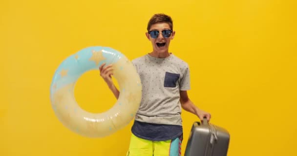  happy teenager in sunglasses stands with a suitcase and an inflatable sea circle on a yellow background - Video