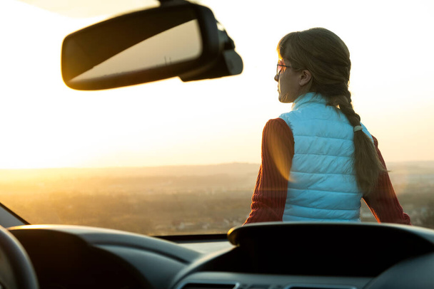 Młoda kobieta stojąca blisko samochodu ciesząca się ciepłym widokiem na zachód słońca. Dziewczyna podróżnik opierając się na masce pojazdu patrząc na horyzoncie wieczornym. - Zdjęcie, obraz