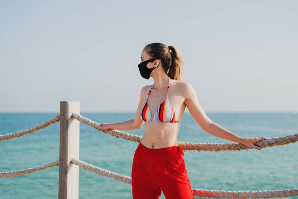 Nuovo concetto normale: giovane donna europea in bikini, pantaloni rossi e maschera protettiva sulla spiaggia pubblica. Vacanza durante la pandemia di coronavirus - Foto, immagini