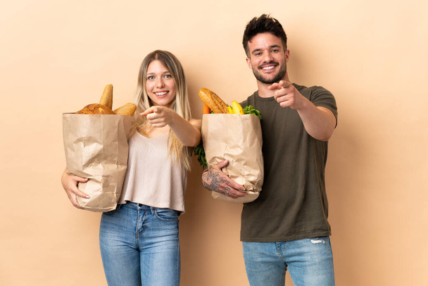 Casal segurando sacos de compras de supermercado sobre fundo isolado aponta dedo em você com uma expressão confiante
 - Foto, Imagem
