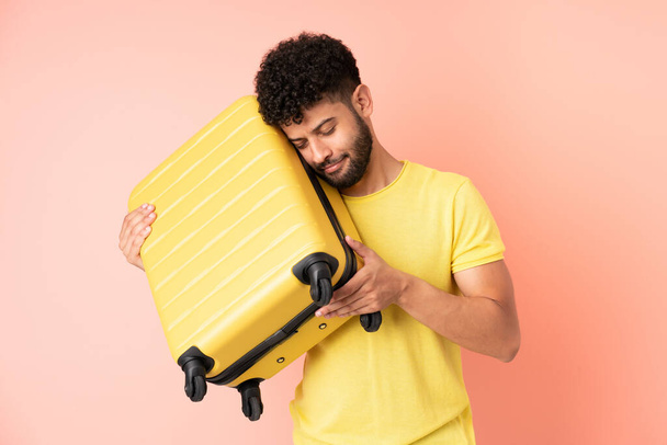 Fiatal marokkói férfi elszigetelt rózsaszín háttér nyaralás utazási bőrönd és boldogtalan - Fotó, kép