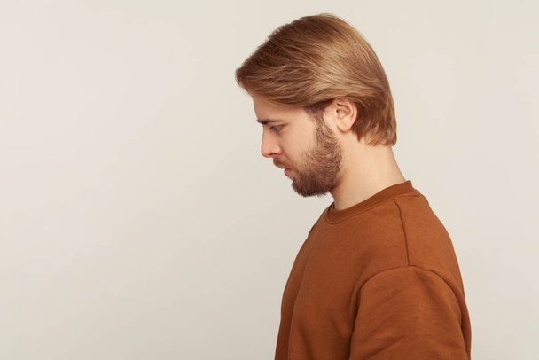 Sadness and resentment emotions. Profile of unhappy bearded man in sweatshirt looking down, bowing head with upset vexed face, depression from life problem. studio shot isolated on gray background - Fotoğraf, Görsel