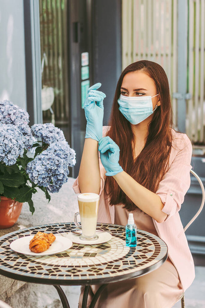 Portrait happy woman in medical face mask put on protective gloves and drink coffee on outdoor terrace in restaurant. Beautiful young girl in protective mask wear medical gloves sit in french cafe - Photo, Image