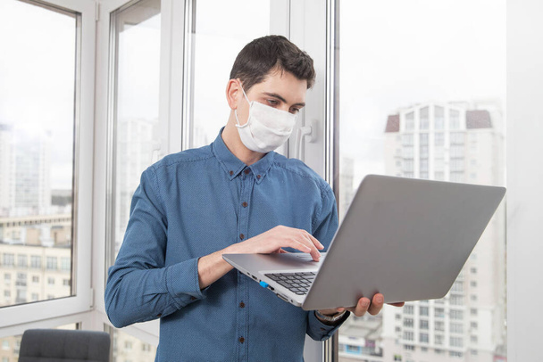 顔の保護マスクの男は、ウイルスの流行中に自宅からコンピュータで動作します。ウイルス感染時の隔離 - 写真・画像